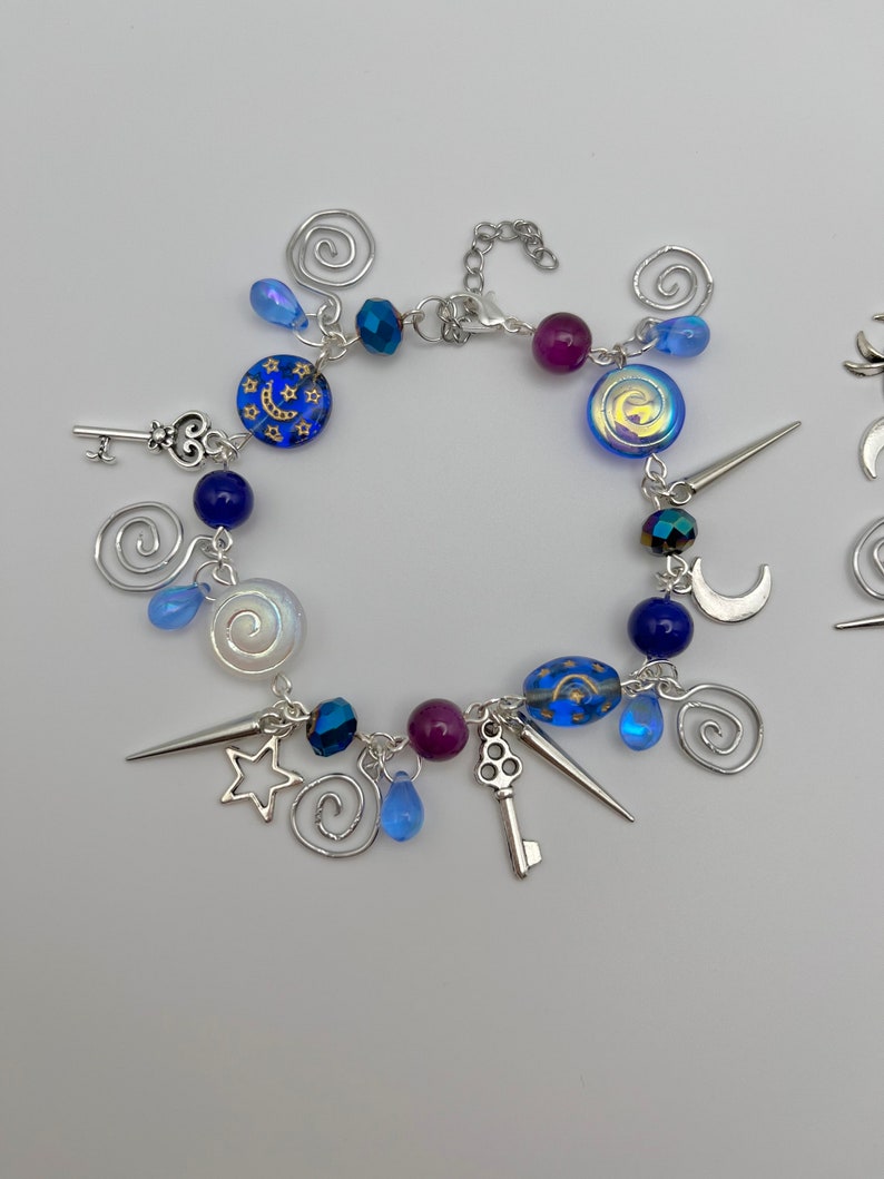 K&ouml;nnte beinhalten: Ein silbernes Armband mit blauen, violetten und wei&szlig;en Perlen und silbernen Anh&auml;ngern, darunter Schl&uuml;ssel, Sterne, Monde und Spiralen.