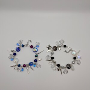 K&ouml;nnte beinhalten: Zwei Charm-Armb&auml;nder mit einer silbernen Kette und Charms. Ein Armband hat blaue, wei&szlig;e und silberne Charms, darunter einen Schl&uuml;ssel, Sterne und Monde. Das andere Armband hat schwarze, wei&szlig;e und silberne Charms, darunter eine Spinne, Sterne und Monde.