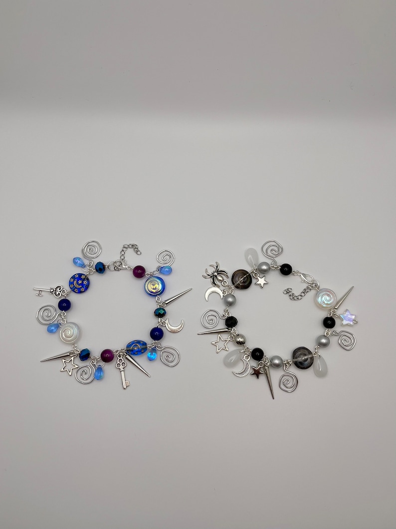 K&ouml;nnte beinhalten: Zwei Charm-Armb&auml;nder mit einer silbernen Kette und Charms. Ein Armband hat blaue, wei&szlig;e und silberne Charms, darunter einen Schl&uuml;ssel, Sterne und Monde. Das andere Armband hat schwarze, wei&szlig;e und silberne Charms, darunter eine Spinne, Sterne und Monde.