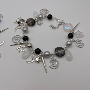 K&ouml;nnte beinhalten: Ein silbernes Armband mit schwarzen und wei&szlig;en Perlen, silbernen Anh&auml;ngern und einem silbernen Kettenverschluss. Die Anh&auml;nger sind Sterne, Monde, Spiralen und eine Spinne.
