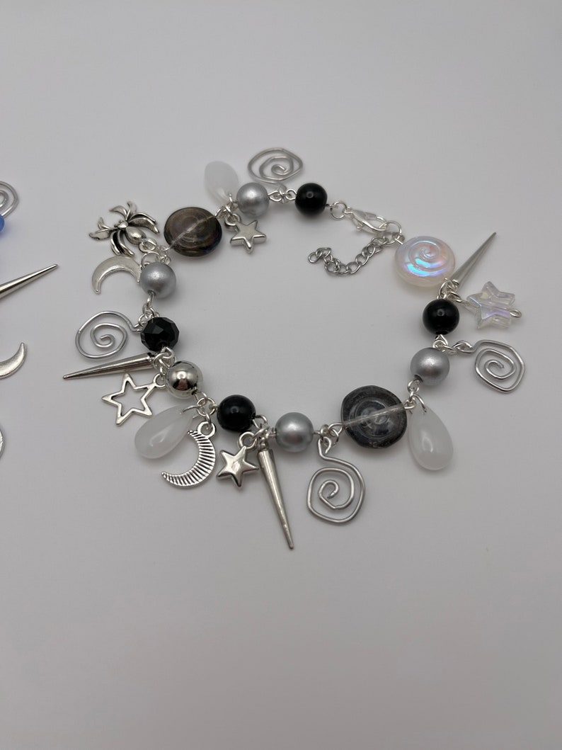 K&ouml;nnte beinhalten: Ein silbernes Armband mit schwarzen und wei&szlig;en Perlen, silbernen Anh&auml;ngern und einem silbernen Kettenverschluss. Die Anh&auml;nger sind Sterne, Monde, Spiralen und eine Spinne.