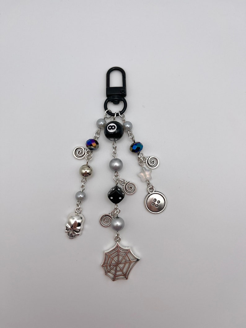 Op de afbeelding: Een zwarte en zilveren sleutelhanger met verschillende bedels, waaronder een schedel, een spinnenweb, een dobbelsteen, een knoop en een zwarte 8-ball.