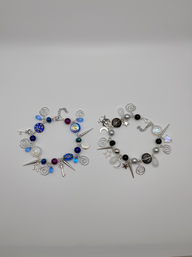 K&ouml;nnte beinhalten: Zwei Charm-Armb&auml;nder mit einer silbernen Kette und Charms. Ein Armband hat blaue und silberne Charms, darunter einen Schl&uuml;ssel, Sterne und Monde. Das andere Armband hat schwarze, silberne und wei&szlig;e Charms, darunter eine Spinne, Sterne und Monde.