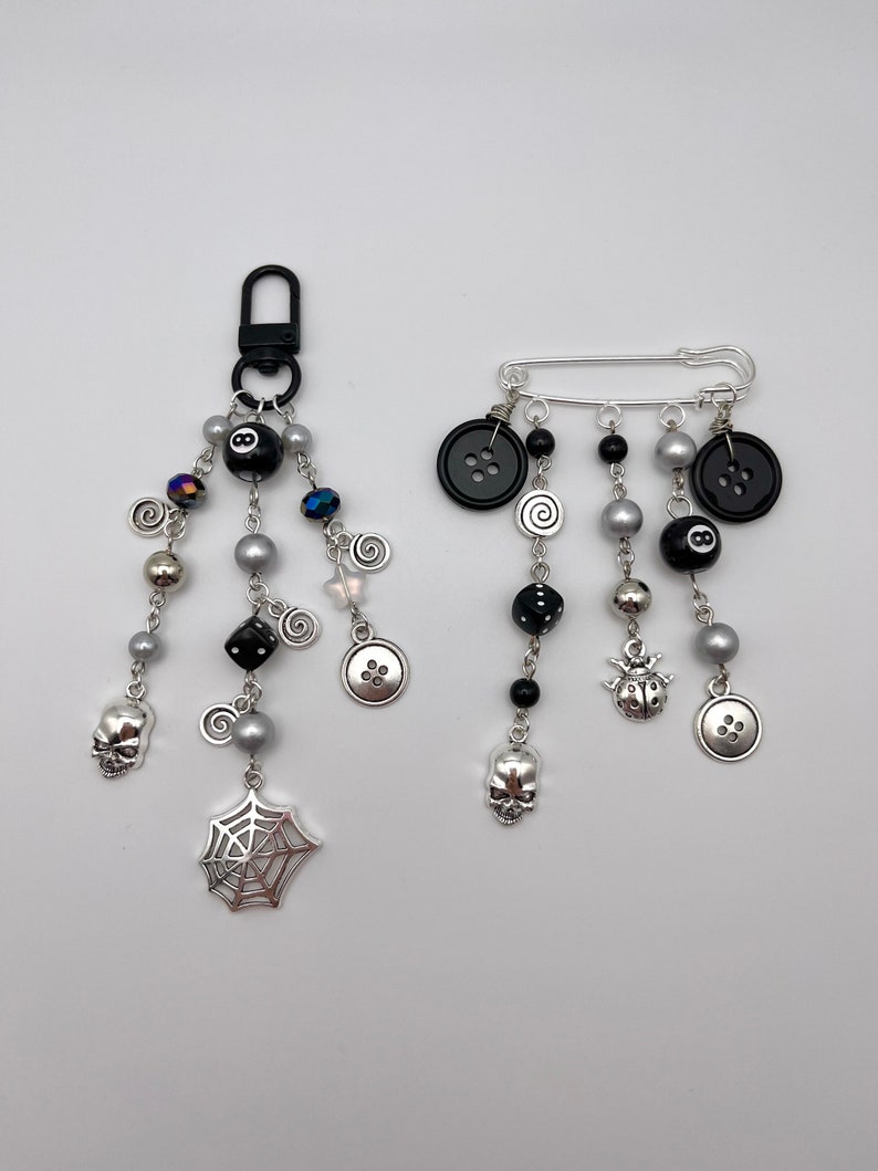Op de afbeelding: Twee zilveren bedelarmbanden met zwarte en zilveren kralen, bedels zijn onder andere een schedel, spinnenweb, dobbelstenen, knopen en een lieveheersbeestje. Een armband heeft een zwarte 8-ball bedel en de andere heeft een veiligheidsspeld sluiting.