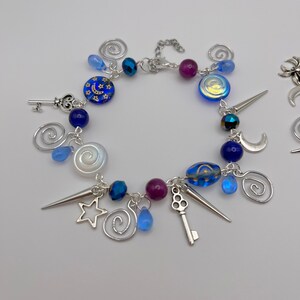 K&ouml;nnte beinhalten: Ein silbernes Armband mit blauen, violetten und wei&szlig;en Glasperlen, silbernen Anh&auml;ngern und einer silbernen Kette. Die Anh&auml;nger umfassen einen Schl&uuml;ssel, einen Stern, einen Halbmond und eine Spirale.