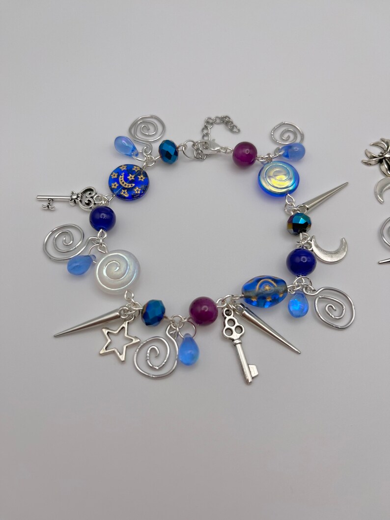 K&ouml;nnte beinhalten: Ein silbernes Armband mit blauen, violetten und wei&szlig;en Glasperlen, silbernen Anh&auml;ngern und einer silbernen Kette. Die Anh&auml;nger umfassen einen Schl&uuml;ssel, einen Stern, einen Halbmond und eine Spirale.