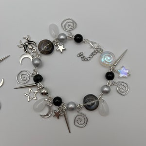 K&ouml;nnte beinhalten: Ein silbernes Armband mit schwarzen und wei&szlig;en Perlen, silbernen Anh&auml;ngern in Form von Sternen, Monden und Spiralen sowie einem silbernen Spinnenanh&auml;nger.