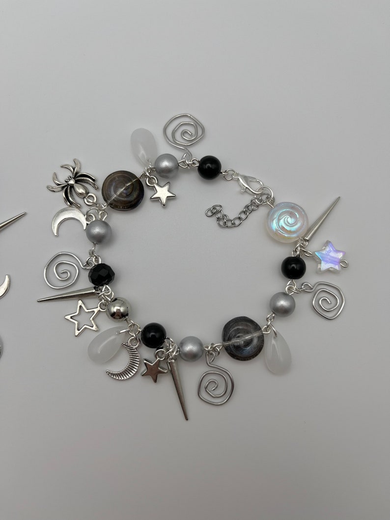 K&ouml;nnte beinhalten: Ein silbernes Armband mit schwarzen und wei&szlig;en Perlen, silbernen Anh&auml;ngern in Form von Sternen, Monden und Spiralen sowie einem silbernen Spinnenanh&auml;nger.