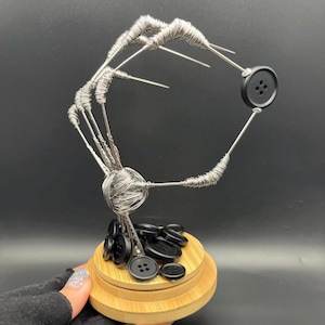 以下が含まれることがあります： 黒いボタンを持った手の銀色のワイヤー彫刻。彫刻は小さな丸い木の台座に取り付けられています。ベースにはいくつかの黒いボタンもあります。ユニークな芸術作品です。