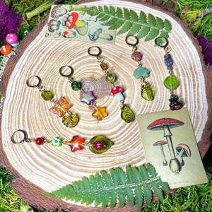 Peut inclure: Cinq marqueurs de points de couture faits à la main en perles de verre et en breloques colorées avec des anneaux dorés. Les breloques comprennent une coccinelle, un champignon, une étoile, une fleur et une feuille. Les marqueurs de points de couture sont présentés sur une surface en bois avec une fougère et une illustration vintage de champignons.