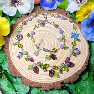 May include: A delicate silver necklace and bracelet set with green, purple, and white glass beads. The beads are shaped like leaves, flowers, and butterflies. The necklace has a charm with a flower design.