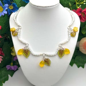 Könnte beinhalten: Eine weiße Perlenkette mit gelben und grünen Blattanhängern. Die Kette ist auf einer weißen Mannequin-Büste ausgestellt.