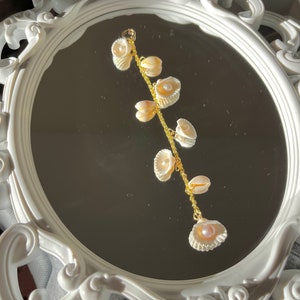 Peut inclure: Un bracelet en chaîne dorée avec une série de petites coquillages blancs attachés. Les coquillages sont disposés en cascade, le plus grand coquillage étant en bas.