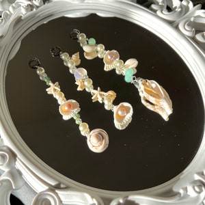May include: Three delicate necklaces featuring seashells, pearls, and green beads. The necklaces are displayed on a white ornate mirror.