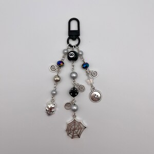 Op de afbeelding: Een zwarte en zilveren sleutelhanger met verschillende bedels, waaronder een spinnenweb, een schedel, een knoop, een dobbelsteen en een zwarte 8-ball. De sleutelhanger is bevestigd aan een zwarte metalen clip.