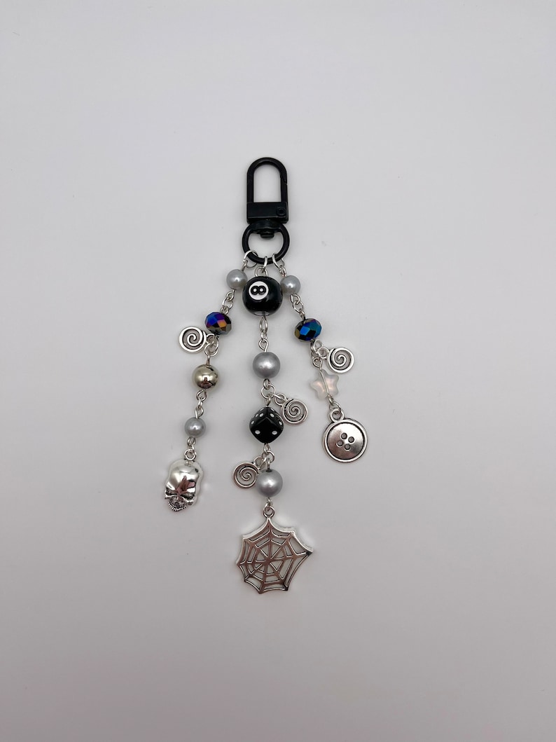 Op de afbeelding: Een zwarte en zilveren sleutelhanger met verschillende bedels, waaronder een spinnenweb, een schedel, een knoop, een dobbelsteen en een zwarte 8-ball. De sleutelhanger is bevestigd aan een zwarte metalen clip.