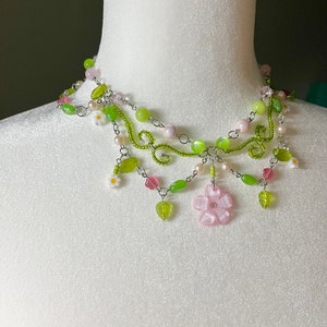 May include: A delicate silver necklace with green and pink beads, featuring a large pink flower charm and smaller white flower accents. The necklace is layered with multiple strands of beads and chain.