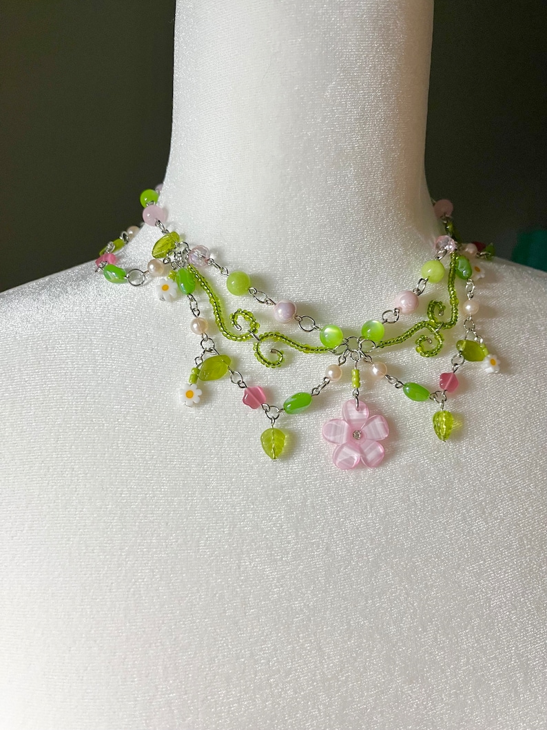 May include: A delicate silver necklace with green and pink beads, featuring a large pink flower charm and smaller white flower accents. The necklace is layered with multiple strands of beads and chain.
