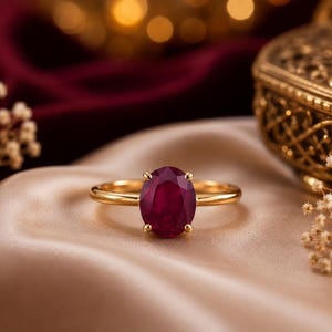 Könnte beinhalten: Ein Goldring mit einem einzelnen, oval geschliffenen, tiefroten Edelstein in einer Vier-Krappen-Fassung. Die Ringschiene ist glatt und poliert und besticht durch ein schlichtes Design, das die leuchtende Farbe und Klarheit des Steins betont.