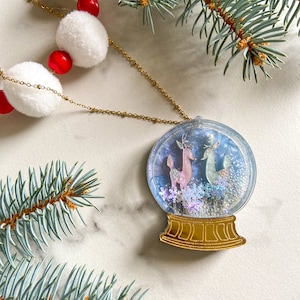Puede incluir: Un collar de cadena dorada con un colgante de bola de nieve. La bola de nieve presenta dos ciervos y copos de nieve. Una guirnalda de pompones blancos y cuentas rojas está en el fondo, junto con ramas de hoja perenne.
