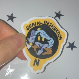 Peut inclure: Un autocollant représentant un personnage de dessin animé aux cheveux bleu clair, aux yeux jaunes et portant un chapeau noir. Le personnage est à l'intérieur d'un cercle jaune avec les mots "SERIAL DESIGNATION" et la lettre "N".