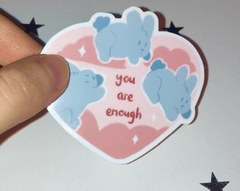 May include: A heart-shaped sticker with a pink and white background, featuring three light blue bunny illustrations. The sticker has the words "you are enough" in red. The sticker is held by a hand.