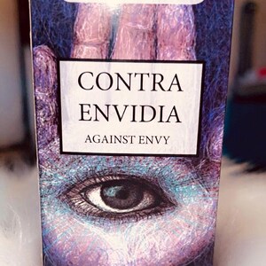 May include: A product box with a mystical design. The box features the text "POWERFUL BATH" at the top and "CONTRA ENVIDIA AGAINST ENVY" in the center. The artwork includes an eye and a hand, with a purple and blue color scheme.