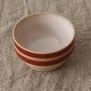 Op de afbeelding: Een kleine, ronde keramische kom met een crèmekleurige buitenkant en een witte binnenkant. Twee horizontale banden van roodbruin omcirkelen de buitenkant van de kom. De kom wordt waarschijnlijk gebruikt voor het serveren van voedsel of als decoratief item.