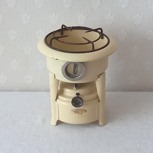 Pode incluir: Um fogareiro de campismo vintage em metal de cor creme, com uma grelha circular no topo, um mostrador central prateado e um pequeno botão de controlo preto na parte frontal. A unidade cilíndrica compacta assenta sobre três pernas.