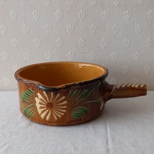 Könnte beinhalten: Ein brauner Keramiktopf mit einem Holzgriff. Der Topf ist mit grünen und weißen Blumenmustern verziert.