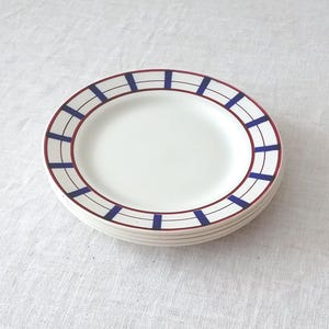 May include: A stack of round white ceramic plates featuring a decorative rim pattern. The rim design consists of thin dark red circular lines intersected by evenly spaced, bold dark blue vertical rectangular bars, creating a grid-like border.