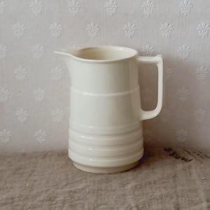 Peut inclure: Un pichet en céramique de couleur crème doté d'une partie supérieure lisse, d'un large bec verseur et d'une anse robuste. La partie inférieure du pichet est ornée de trois bandes horizontales en relief entourant le corps cylindrique.