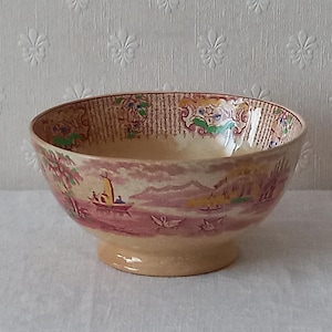 Może przedstawiać: Wazon ceramiczny w stylu vintage z motywem krajobrazu. Wazon ma kremową podstawę i obrzeże ze szczegółowym wzorem. Na zewnątrz widnieje scena z łodziami, górami oraz różowo-kremową paletą barw.