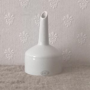 Può includere: Un imbuto in ceramica bianca con una base larga e un beccuccio stretto e affusolato. La superficie esterna è liscia e lucida. Vicino al bordo inferiore, è presente un piccolo timbro ovale contenente le lettere A G D in un carattere scuro.