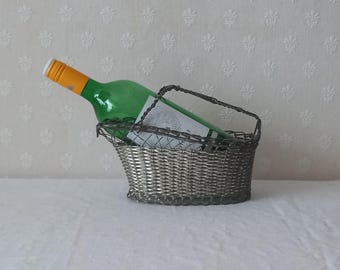 Puede incluir: Una botella de vino verde con tapón naranja descansa en una cesta de alambre plateado con asa. La cesta tiene un diseño tejido y está sobre una superficie blanca. La botella está parcialmente llena de líquido.