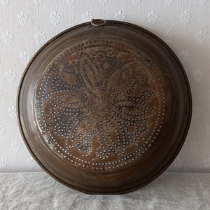 Puede incluir: Un colador de cobre vintage con un patrón floral decorativo. El colador es redondo y tiene un asa en la parte superior.