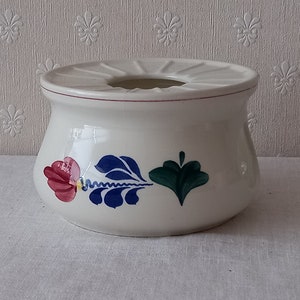 Puede incluir: Olla de cerámica blanca con un diseño floral rojo, azul y verde. La olla tiene una parte superior ancha y plana con un agujero en el centro.