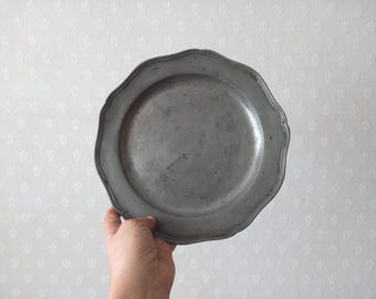 Puede incluir: Un plato redondo de peltre con un acabado metálico gris mate y un borde festoneado liso. La superficie del plato muestra sutiles signos de desgaste y una pátina natural, con un centro ancho y plano y un diseño de borde ligeramente curvado.