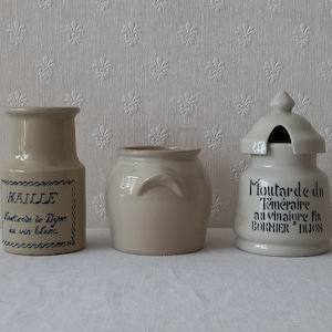 Op de afbeelding: Drie beige keramische containers. De container aan de linkerkant heeft een label met blauwe tekst met de tekst "MAILLE Moutarde de Dijon au vin blanc". De middelste container is een kleine pot met handvatten. De container aan de rechterkant heeft een deksel en de tekst "Moutarde du Téméraire au vinaigre fin BORNIER DIJON."