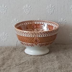 Op de afbeelding: Een decoratieve keramische kom met een witte voet. De kom heeft een bruin en wit geometrisch patroon met ruit- en vierkantvormen. De rand heeft een herhalend gestippeld ontwerp. De kom wordt waarschijnlijk gebruikt voor het serveren van voedsel of als decoratief item.