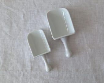 May include: Two white ceramic scoops with smooth, rounded handles and deep, rectangular bowls. The scoops are glossy and feature a clean, minimalist design with a uniform white finish throughout the entire structure of both kitchen utensils.