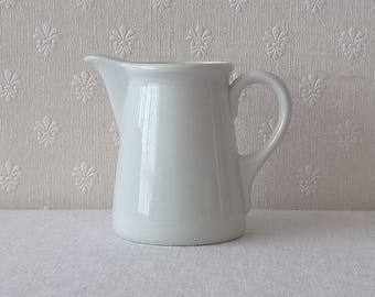 May include: A plain white ceramic creamer pitcher with a smooth, glossy finish. The pitcher features a wide, rounded body, a curved handle on the right side, and a prominent pouring spout on the left side, creating a simple and functional design.