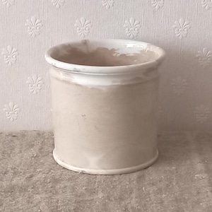 May include: A cylindrical ceramic vessel featuring a smooth, light beige and white marbled finish. The container has a wide, open top with a slightly flared rim and a flat base, showcasing a consistent, neutral-toned swirling pattern throughout.