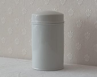 May include: A cylindrical light gray storage canister with a smooth, solid finish and a matching fitted lid. The container features a simple, vertical design with a rounded top edge, providing a clean and uniform appearance for household storage.