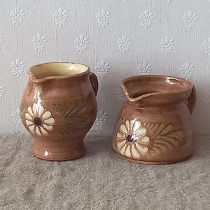 Pode incluir: Dois jarros de cerâmica vidrada castanha com um motivo de margarida em cor creme pintado à mão, com um centro escuro e detalhes de folhas verdes. Cada jarro tem um corpo arredondado, um pequeno bico vertedor e uma pega lateral brilhante.
