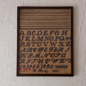 May include: A framed sampler with a brown wooden frame. The sampler is made of beige fabric with black embroidery of the alphabet, numbers 1-9 and 0, and the text "B. Macy 1917".