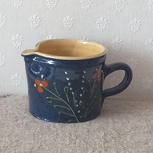 Op de afbeelding: Een donkerblauwe keramieken melkkannetje met een lichtbruine binnenkant en één handvat. De buitenkant is voorzien van een handgeschilderd bloemmotief met oranje bloemblaadjes, groene stengels en kleine witte stippen in verticale lijnen.