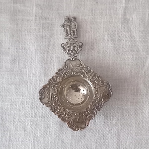 Op de afbeelding: Een theezeef van zilverkleurig metaal met een ruitvormige kom voorzien van gedetailleerd reliëfwerk met krullen en landschappen. Het handvat is bekroond met twee kleine staande figuren en een sierlijk ornament boven de kom.