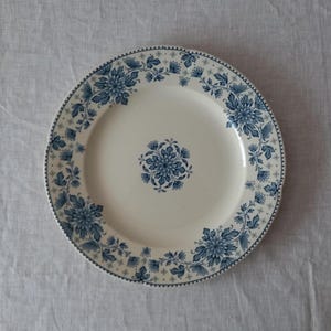 Op de afbeelding: Witte keramische plaat met een blauw bloemmotief. De plaat heeft een centraal bloemmotief en een rand van kleinere bloemen en bladeren. De rand heeft een geschulpte rand. De plaat staat tegen een neutrale achtergrond.