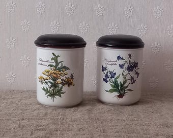 May include: Two white ceramic canisters with black lids. One canister features a botanical illustration of yellow flowers and green leaves, labeled "Chrysanthemum parthenium". The other displays blue bellflowers, labeled "Campanula rotundifolia".